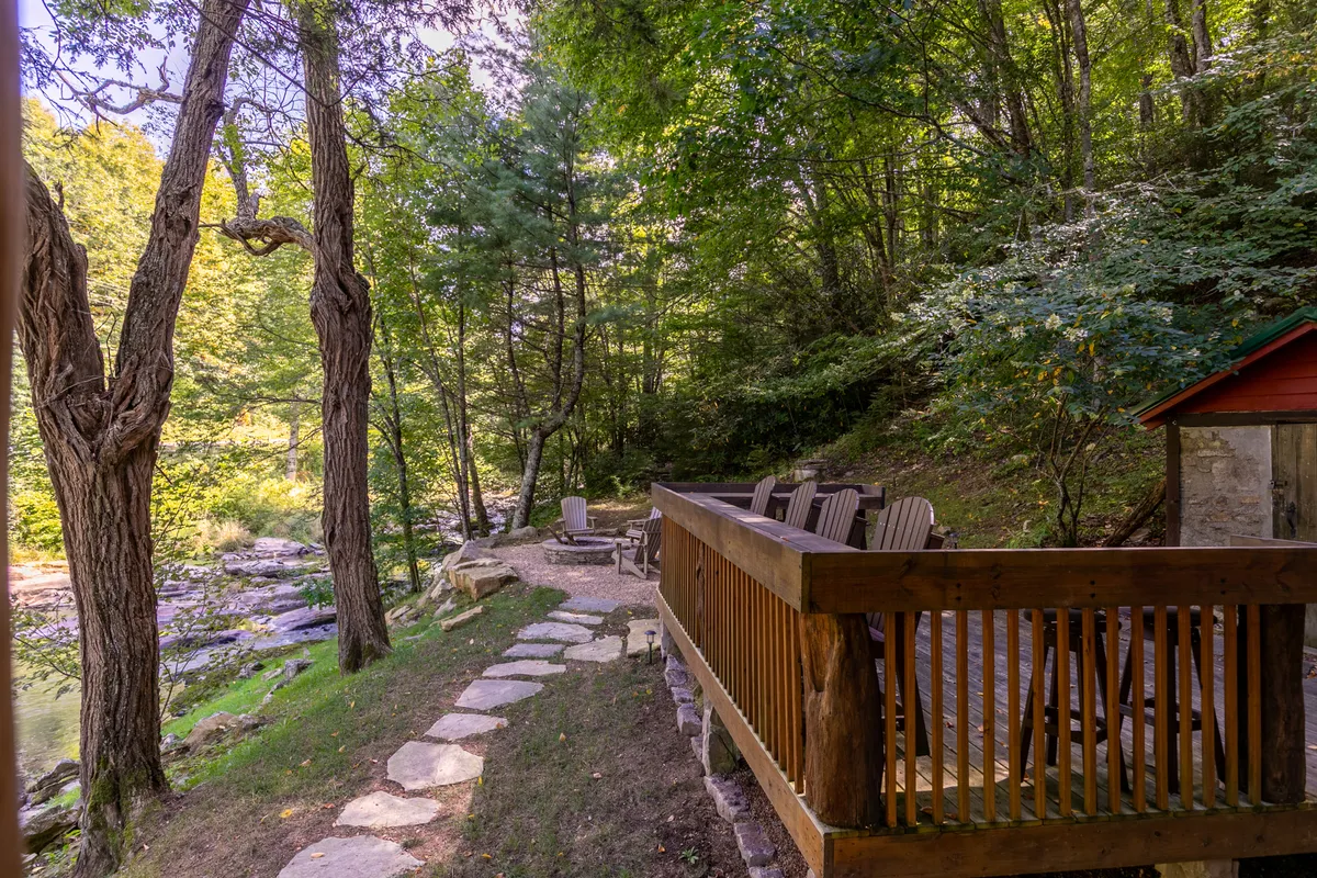 A welcoming exterior scene featuring a wooden deck surrounded by mature trees, with Adirondack chairs positioned around a stone fire pit. The setting exudes serenity and invites outdoor gatherings, relaxation, and the enjoyment of shared amenities like the fire pit and deck space. The natural woodland backdrop highlights the private-yet-social atmosphere of the property.