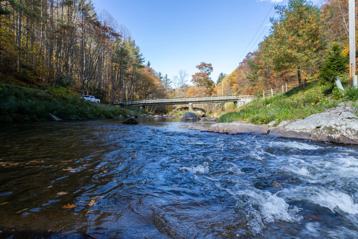 A gently flowing river winds past a sturdy bridge, framed by vibrant autumn foliage and scattered rocks. The peaceful scene hints at prime fishing spots, quiet riverside walks, and wildlife observation. The rich colors add seasonal atmosphere, emphasizing the connection to mountain nature and the property's private trail network.