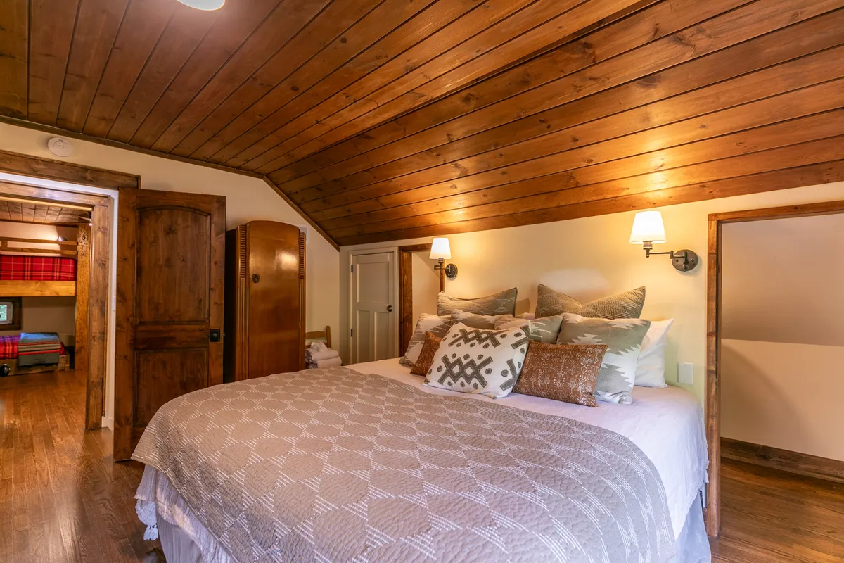A spacious bedroom with a large, comfortable bed adorned with plush pillows, a wooden ceiling, and soft lighting from wall sconces. The atmosphere is calm and quiet, evoking restful nights and peaceful mornings.