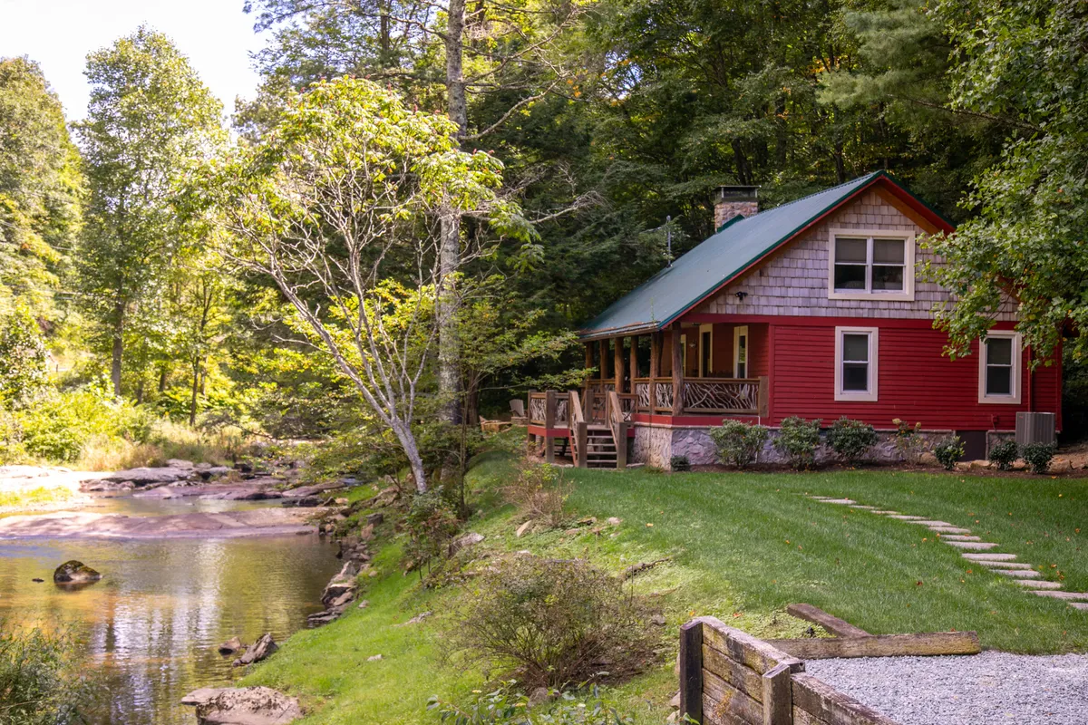 This image showcases a red cabin perched on the bank of a flowing river, surrounded by verdant trees and a tranquil stone pathway. The porch of the cabin offers a front-row seat to the river, perfect for fishing, sipping morning coffee, or relaxing as the sun sets. The overall mood is peaceful and inviting, emphasizing a deep connection to the river and the natural environment.