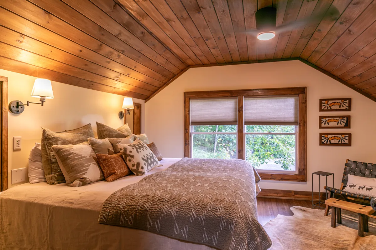 A welcoming bedroom featuring a cozy bed with decorative pillows set against a backdrop of large windows, a wooden ceiling, and tasteful artwork on the walls. The room is washed in natural light, offering views of the surrounding landscape and a sense of warmth.