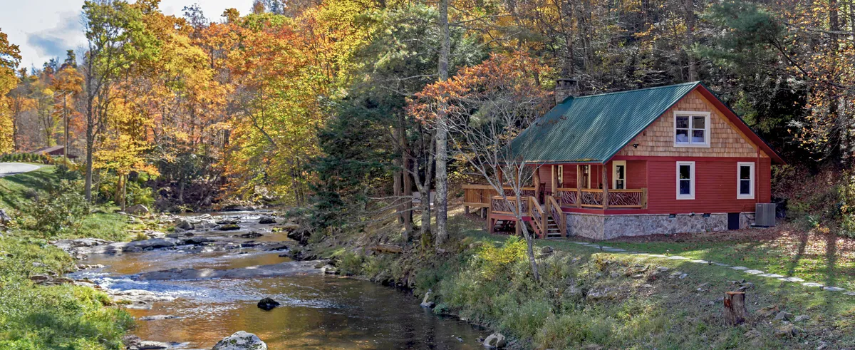 A colorful autumn landscape featuring a cabin by a stream, surrounded by vibrant trees. The scene invites fishing, hiking, and relaxing by the water—seasonal activities that encourage families to gather, celebrate, and enjoy nature together.