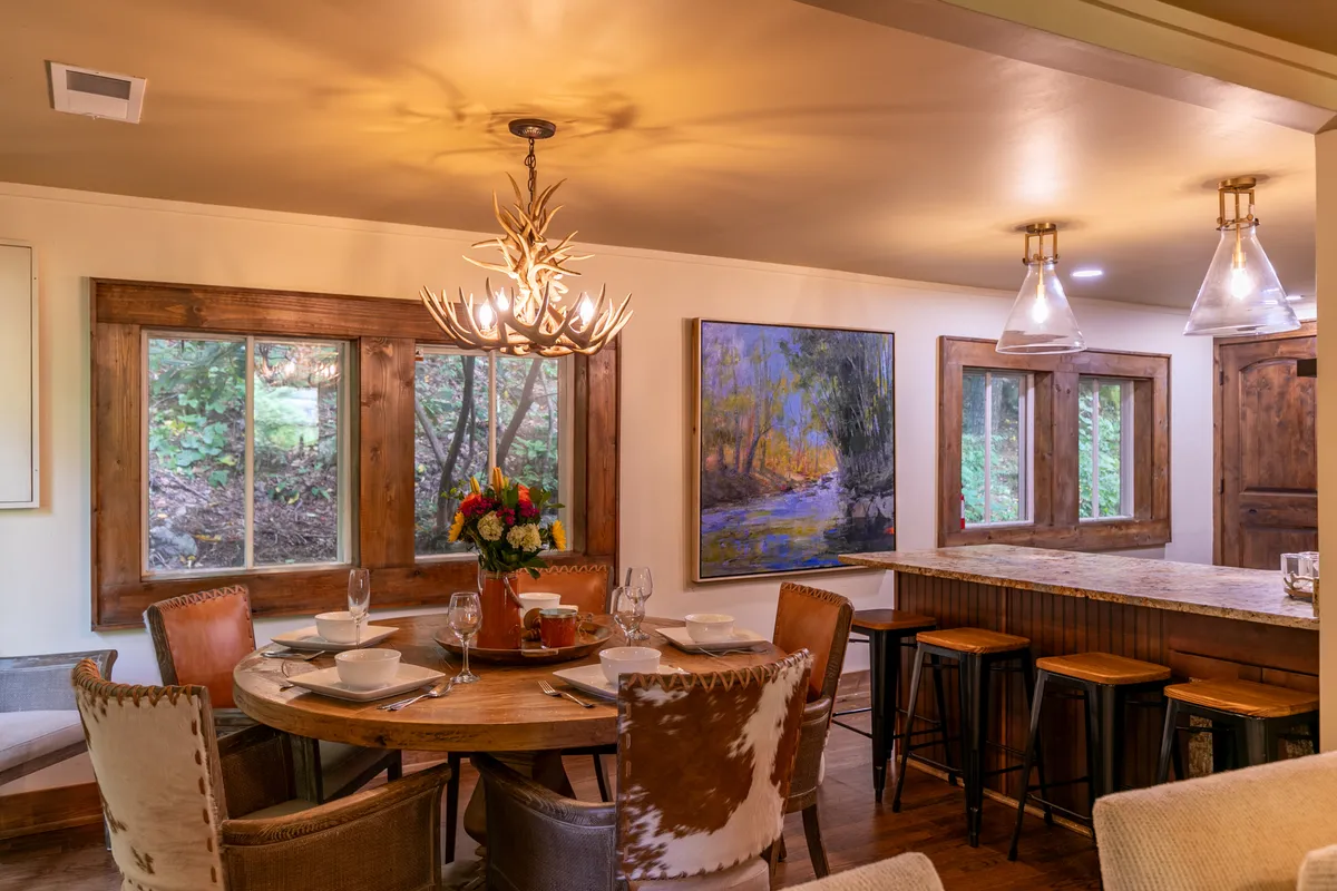 A large wooden dining table set beneath a chandelier, with a painting adorning the wall and a glimpse of a bar area nearby. The space is arranged to welcome groups for meals, conversation, and celebration.