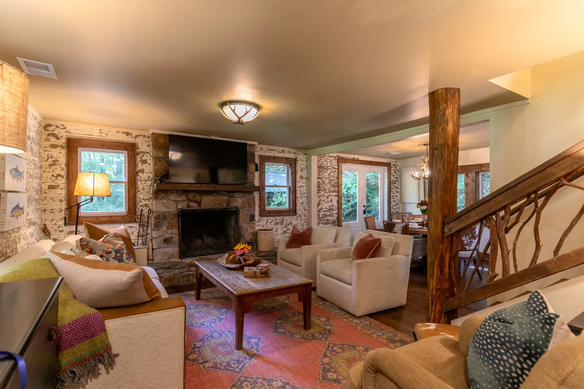 A cozy living room featuring a prominent stone fireplace, a comfortable seating area with plush chairs and a sofa, a coffee table adorned with decorative items, and large windows that let in ample natural light. The view through the windows hints at the beauty of the surrounding river and greenery, while vintage details like decorative pillows and classic finishes evoke 1930s charm.