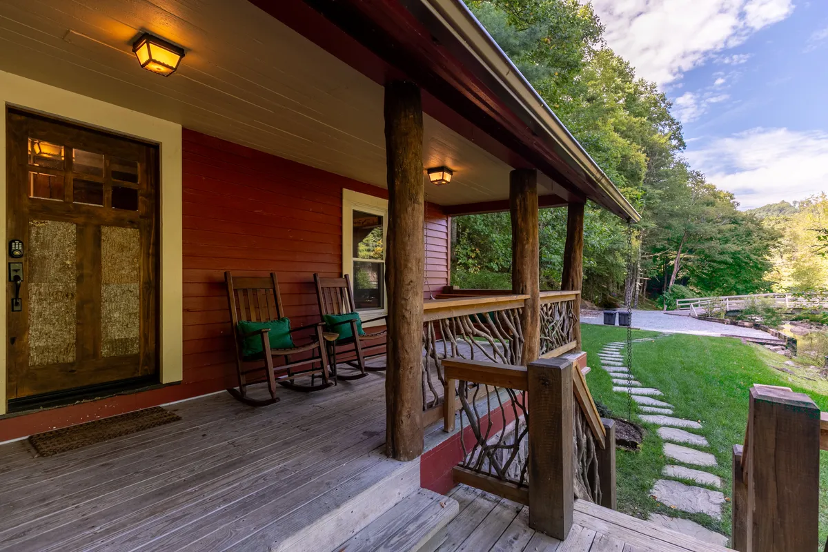A welcoming front porch adorned with rocking chairs and a sturdy wooden railing, set against a backdrop of lush greenery and a winding pathway. The porch is positioned to take in sunset views, offering an inviting transition space between the interior living room and the river just beyond.