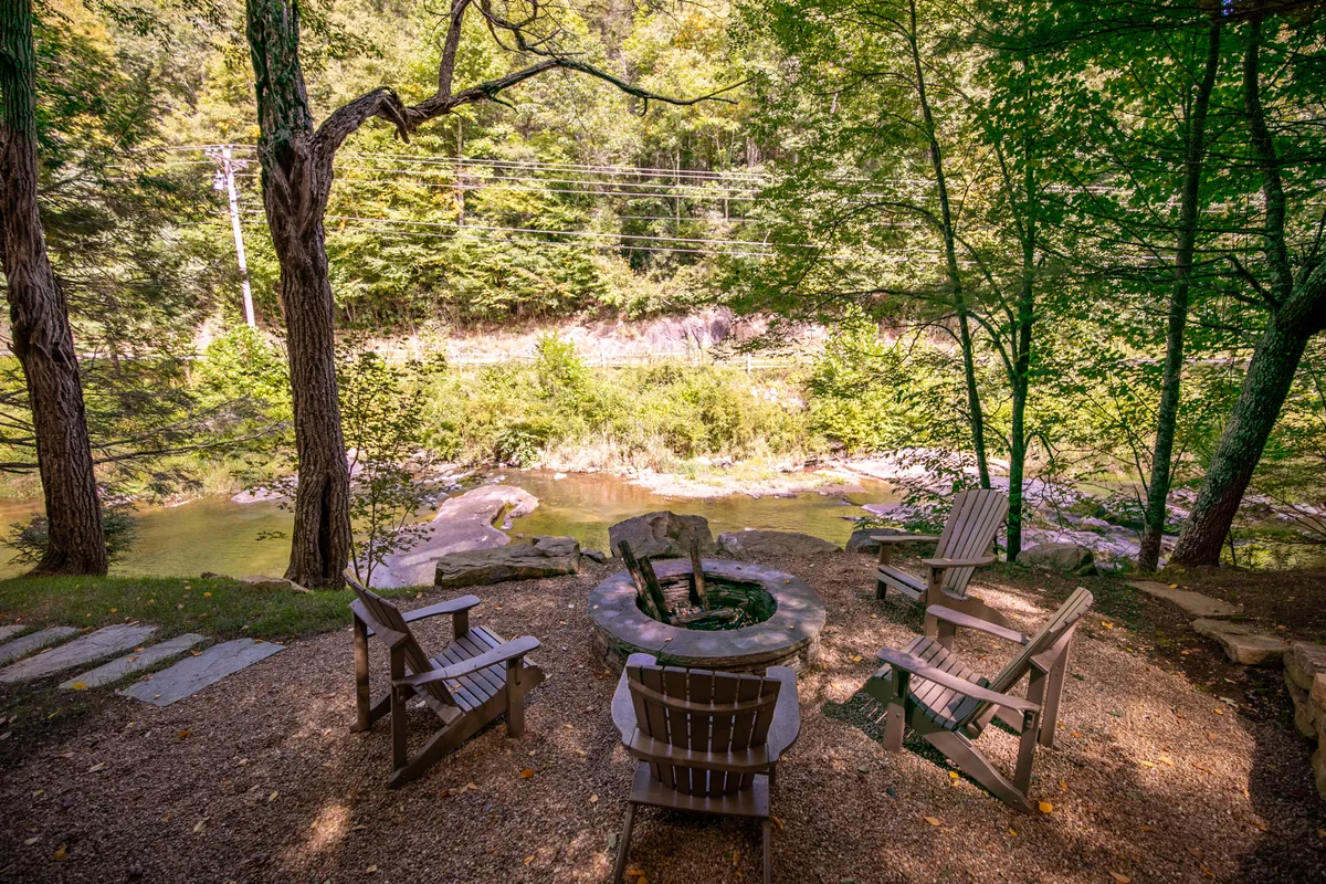 This image features a cozy fire pit surrounded by Adirondack chairs overlooking a tranquil river, set against a backdrop of lush trees. The soft evening light hints at sunset, evoking a sense of warmth and seclusion, ideal for intimate gatherings or quiet romantic moments under the stars.