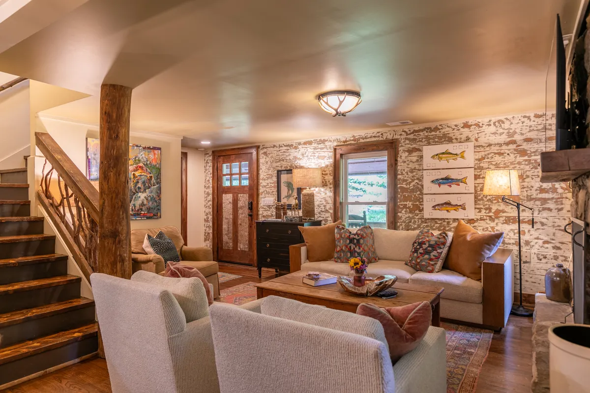 A warm, inviting living area with a plush sofa adorned with cushions, a decorative coffee table, art-filled walls, and a wooden staircase. The space feels relaxed and communal, suitable for transitioning between outdoor adventures and cozy indoor moments.
