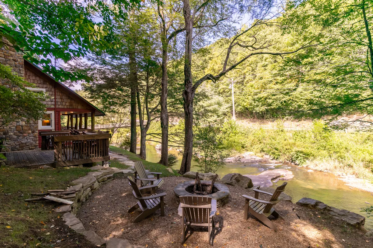 This image captures an exterior scene featuring a cozy fire pit surrounded by wooden chairs, with a picturesque river, trees, and part of the cabin in view. The setting invites guests to enjoy nature, gather by the fire, and take in the tranquil atmosphere that is appealing year-round.