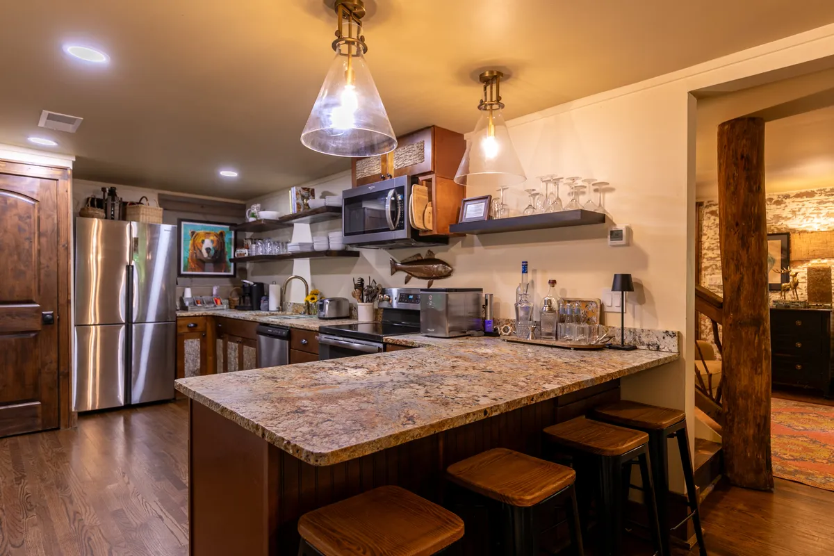 A spacious kitchen area featuring a granite countertop bar with wooden stools, stainless steel appliances, open shelving with glassware, and an inviting, open-plan design that connects easily to the dining space.
