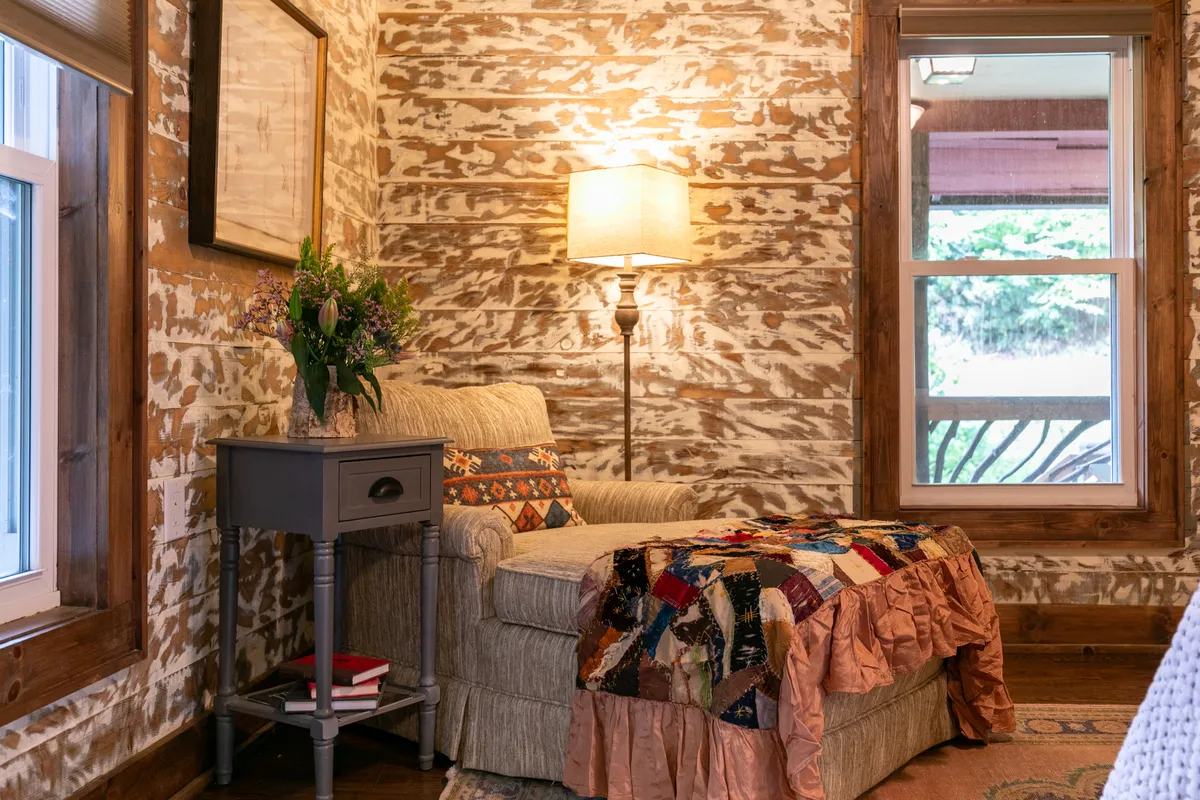 A homey corner of the living room featuring an overstuffed armchair draped with a quilt, a classic lamp, a side table with a floral arrangement, and vintage woodwork. Sunlight streams in through nearby windows, creating an inviting spot for reading, relaxing, or savoring a quiet cup of coffee while gazing at the riverside view.