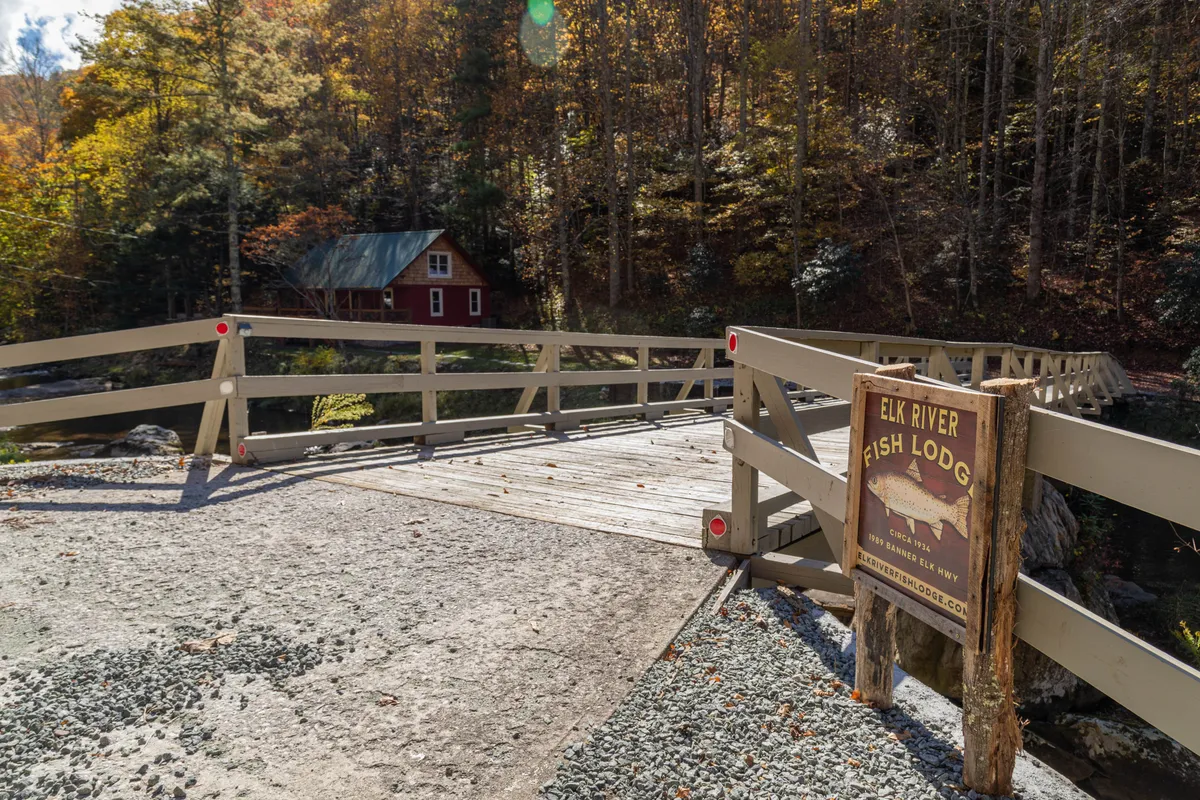 A charming red lodge sits beside a wooden bridge that spans the river, framed by lush trees and a rustic signboard. The riverside location is highlighted by soft sunset light, suggesting quiet moments spent watching the water or heading out for a hike or fishing trip. The bridge and river setting reinforce the exclusive river access and the quiet, historic character of the property.
