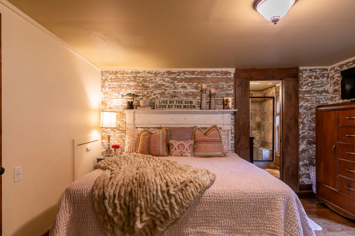 A serene bedroom featuring a bed with fluffy, decorative pillows, a cozy blanket, a wall sign, and wooden accent pieces. The soft lighting and carefully chosen details create a comforting and restorative environment.