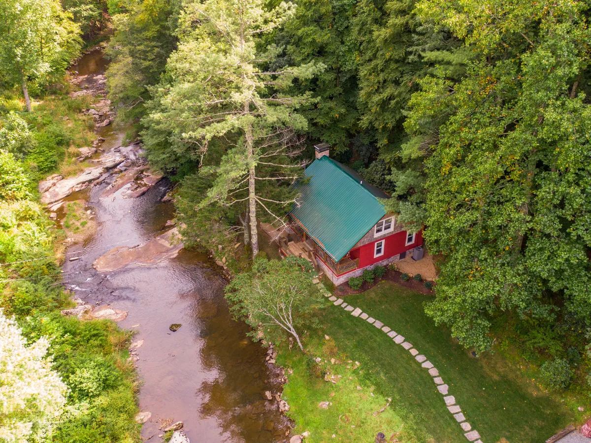 The image displays a charming, renovated red house nestled along the banks of a sparkling river, surrounded by lush trees and a peaceful natural setting. A winding pathway leads guests toward the water, inviting exploration and outdoor activities.